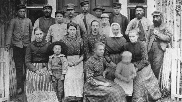 Husmannsfolk og arbeidere på Majer, ca. 1890 - Fotografert foran hovedinngangen i Nistugua på Majer. I bakre rekke er nr. 2 Håken Nyborg, husmann i Esselstuen, nr. 4 er Martin Majerskogen, nr. 5 Ole Kalvsvea, nr. 7 Ole Jakob Skogheim og nr. 8 het Haraldsbakken. Nr. 2 i andre rekke er Hans Nyborg. Navnene på de øvrige er ukjent. Majer var en relativt stor gard, og i 1865 var det fem husmannsplasser under garden. – Fotograf: Ukjent. Foto: Mjøsmuseet / R 80/003/13. Gruppe på 18 voksne og barn foran et hus