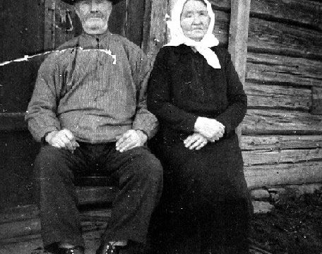 Husmannsfolk i Korsvollhagen, 1925 - Kristian Mathiassen Korsvollhagen (f. 1865) og kona Inge Maria Kristiansdatter (f. 1861), fotografert foran stua på plassen Korsvollhagen under Aslakrud i Kolbu ca. 1925. Inge Maria var da blitt blind. På husmannsplassen hadde de ei ku, en gris og noen høner, og ca. fem mål jord. – Fotograf: Ukjent. Foto: Mjøsmuseet / R 79/025/06. Sittende eldre mann og kvinne