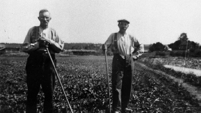 Husmenn på Rogneby, Østre Toten - 1. Peder Anton Lauritsen Gaarderhagen, f. 9. mars 1875, d. 24. oktober 1961. 2. Bernt Martinsen Møllerhaugen, f. 11. april 1882, d. 27. desember 1952. Bildet er tatt før 1950 på Rogneby i Østre Toten. – Fotograf: Ukjent. Foto: Mjøsmuseet / R 90/031/06. To menn som står med verktøy på et jorde