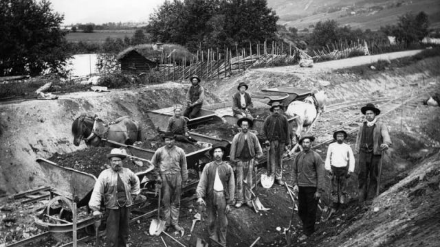 Jernbanen gjennom Gudbrandsdalen ble bygd i midten av 1890-åra, og skapte mange nye arbeidsplasser for husmenn. Bildet viser H. H. Kirketeigens arbeidslag ved Kvam i Gudbrandsdalen. – (Foto: Hans H. Lie, Maihaugen - SS-HHL-03048) 11 menn ved jernbanebygging poserer med verktøy og fulle lass med jord kjørt med hest og kjerre
