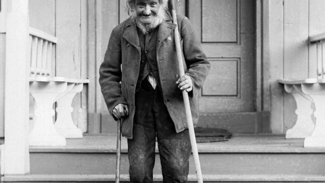 Ole Knipen, husmann under Veikle i Kvam i Nord-Fron, fotografert i juni 1897. – (Foto: Hans H. Lie, Maihaugen - SS-HHL-03581) Eldre mann med rake og gåstokk foran dør