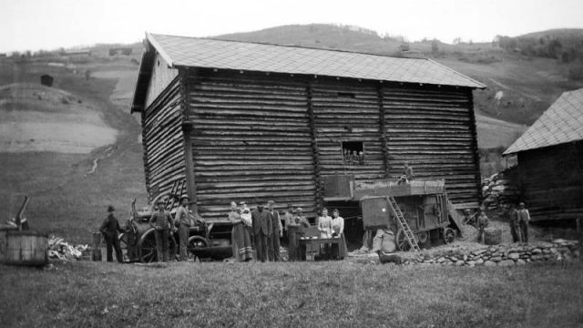 Tresking med treskeverk som blir drevet av lokomobil. Brandvol i Fron 1901 – (Foto: Hans H. Lie, Maihaugen - SS-HHL-C-00003) Flere mennesker står rundt høyt trebygg med maskineri på siden av bygget