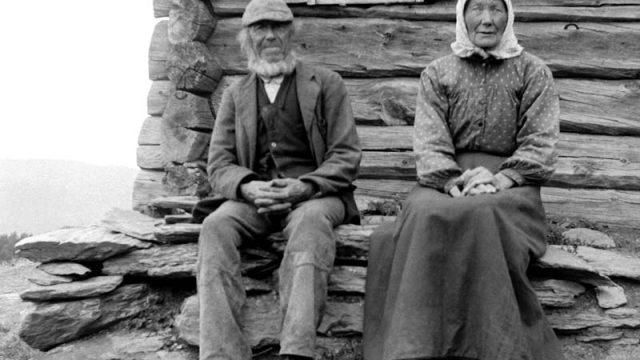 To gamle husmannsfolk fotografert utenfor stua si. – (Foto: Hans H. Lie, Maihaugen - SS-HHL-C-00038) Gammel mann og kvinne sitter foran trebygg