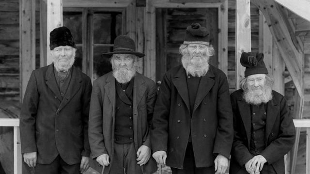 Fire brødre fra Skjåk fotografert rundt 1905. Bønder og husmenn hadde ofte samme opphav, og innenfor samme barneflokken kunne noen bli sjøleiende bønder og andre husmenn. Av disse fire brødrene var det både et par gårdbrukere og en husmann. Fra venstre: Peder Aurstad f. 1832, Ola Bakke f. 1827, Ola Rudsar f. 1829 og Erik Nørstenesstuen f. 1824. – (Foto: Kristian Stakston, Maihaugen - SS-KS-A-0767) Fire eldre menn, tre av dem med stokker, poserer foran et hus dør til første etasje og med trapp til andre etasje